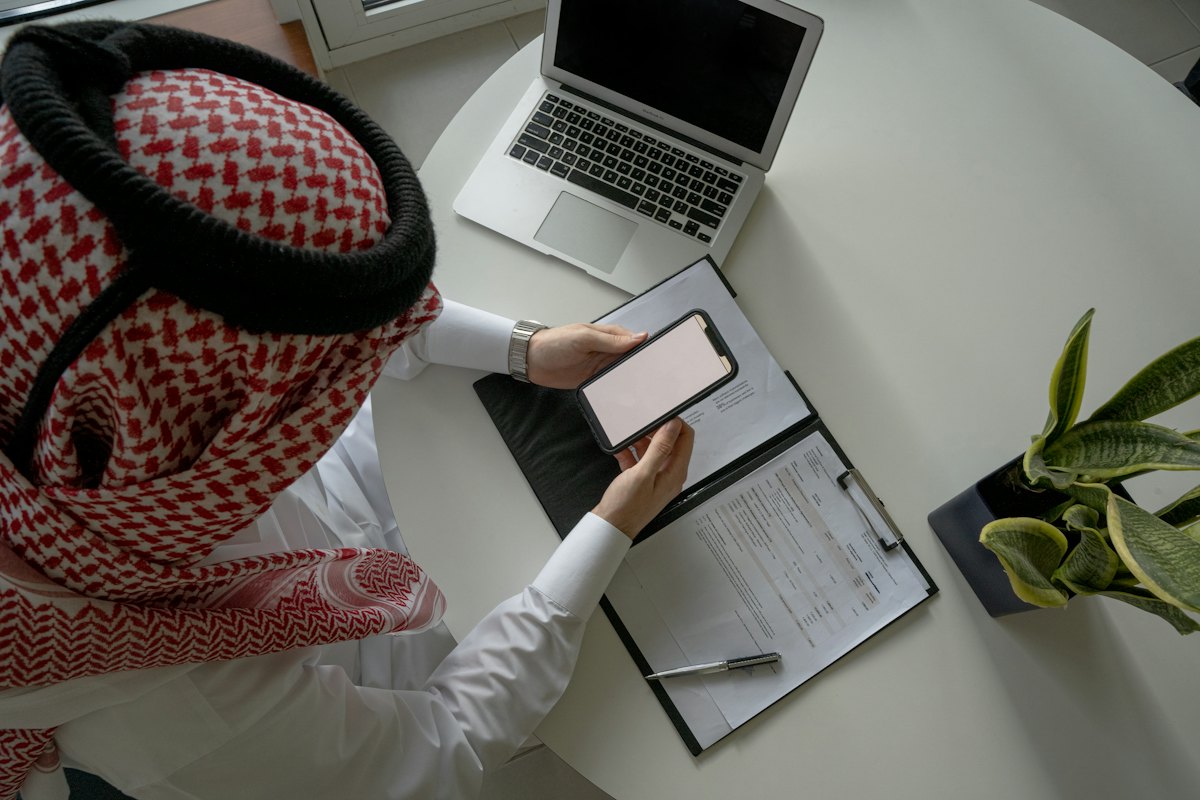 A professional in a ghutra reviewing documents on a phone and laptop
